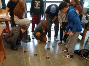 Students stand around a chalk track that has 5 roughly straight lanes and car models racing down them propelled by rear propellers. 