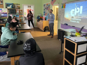 Two adults stand in front a of classroom seated at lab tables. On the projection screen is an image of high power transmission lines and the text "CPI CONSUMERS POWER INC."