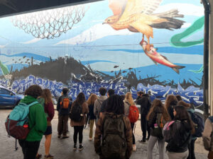 Students with backpacks look at a mural on a wall that has an osprey with a trout flying over a black jagged landscape with fish swimming over it and blue graffiti text in a jumble. 