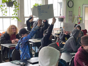 Students in a classroom. Several hold up whiteboards. The whiteboard in the front reads, "Power, Solar, Hydro, Generators, renewable energy, nuclear