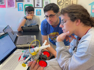 Students sit in front of laptops. One student is holding a model car that has several syringes on top and alligator clip wiring.