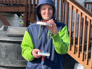 Boy holds a marine wave generator model made from a float, foam, fishing line, a plastic tube wrapped in copper wire, a magnetic hook and magnet with suction cup bottom.