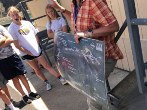 An adult holds a posterboard with a structure showing it to students standing opposite. Text that can be read at the bottom says, " Water Withdrawal" The PGE logo is in the upper right corner.