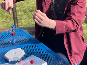 Outside on a blue table a student builds a marine wave energy generator model using a plastic tube sitting on a suction cup to the table. The tube is wrapped in red copper wire. the student uses fishing line to control a magnetic hook with magnets in the tube. Foam rests on the table.