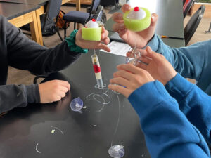 3 sets of hands hold 2 marine wave energy generator models. 2 hands hold green foam with fishing floats in the middle. Fishing line is connected to the foam extending into plastic tubes, 1 tube is held in 2 hands and can't be clearly seen. The other tube is suspended above a black lab table, it is wrapped in red copper wire. 2 clear suction cups are on the table.