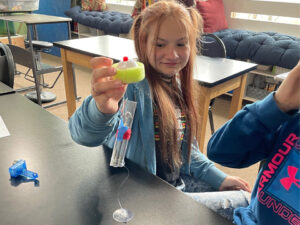 A girl holds a marine wave energy generator model by the float incased in foam. Below is a plastic tube with copper wire wrapped around it. Inside the tube a magnetic hook can be seen. The tube has fishline connecting it to a suction cup stuck to a lab table. 