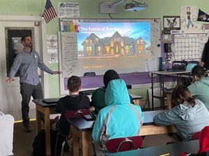 Man presents to a classroom seated at lab tables. On the screen is a projection of a house with the title "The Future of Energy" at the bottom of the slide it reads "Building Electrification"