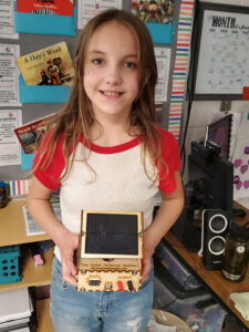 A girl holds a wooden kit that has a solar module on top of a box labeled "The Solar Science Station" The front of the box reads "Volts" with a 3 digit LED screen below it, "USB" with a usb port, and "OUTPUT" with a red and black banana clip plug.