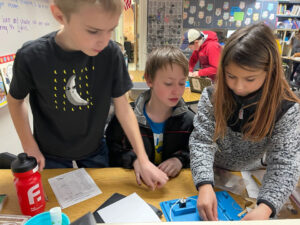 3 kids work at a table with a blue plastic tray that has a motor with wires. 