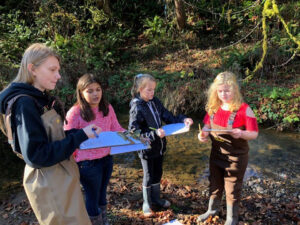 3 girls stand with a young woman in a stream wearing water boots and carrying clipboards.