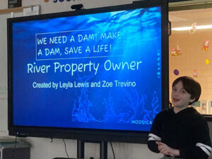 2 girls present using a digital screen the screen reads, "The Disadvantages of not Having a Dam. Some disadvantages of not having a dam are: There might be more floods, Crops might be damaged in floods, We won't have more electricity, But to prevent these, we could: Have a watch tower near the lake with shifting guard rotations, to alert us of any potential danger, We can set up wind turbines and solar panels, connect them to a power plant or two to generate electricity that way.