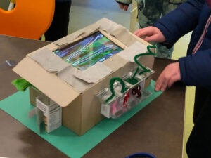 A cardboard box with a window in the top lid. Plastic with pipe cleaners on the long edge and a cardboard door to the front. 