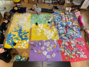 Kids lay around several large sheets of colored paper covered in different types of plastics. The plastics are sorted with wrappers to the right, containers to the middle, bags and gloves to the left.